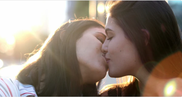 Two women are sharing a kiss outdoors in warm, golden sunlight. The close-up shot captures an intimate moment, with soft lens flare and a blurred background enhancing the romantic atmosphere.