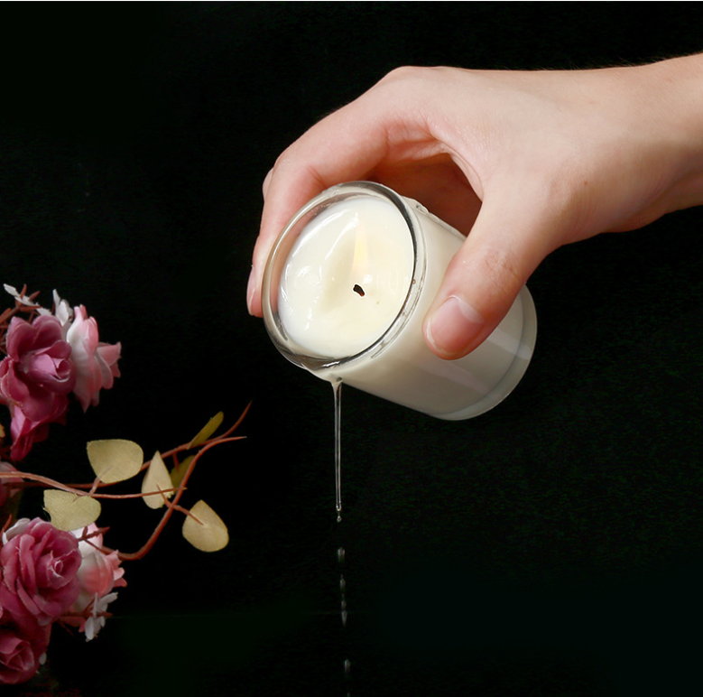 Hand holding Aromatherapy Massage Candle for sex, with coconut wax melting, surrounded by flowers.